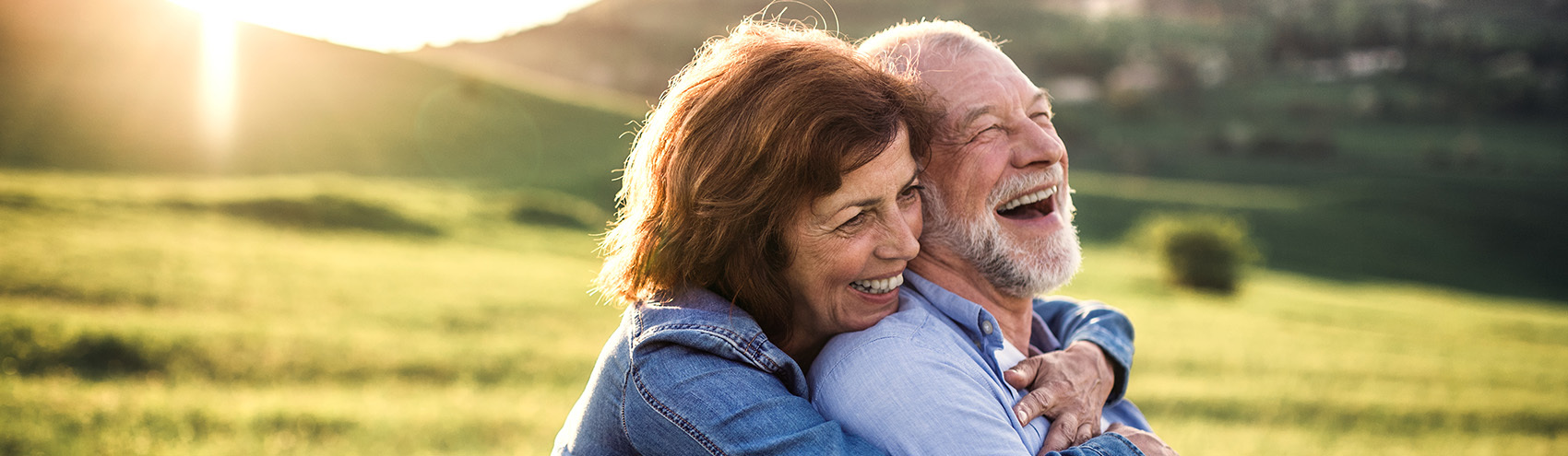 Older couple embracing and laughing, showing off their dental implants in a green field in Austin, TX